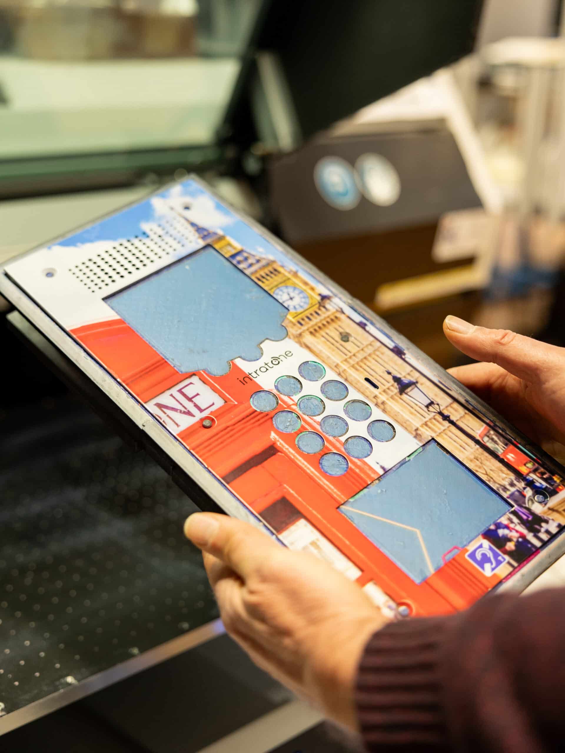 Personne tenant un interphone Intratone illustrée avec un visuel urbain, un clavier numérique circulaire et des éléments graphiques colorés